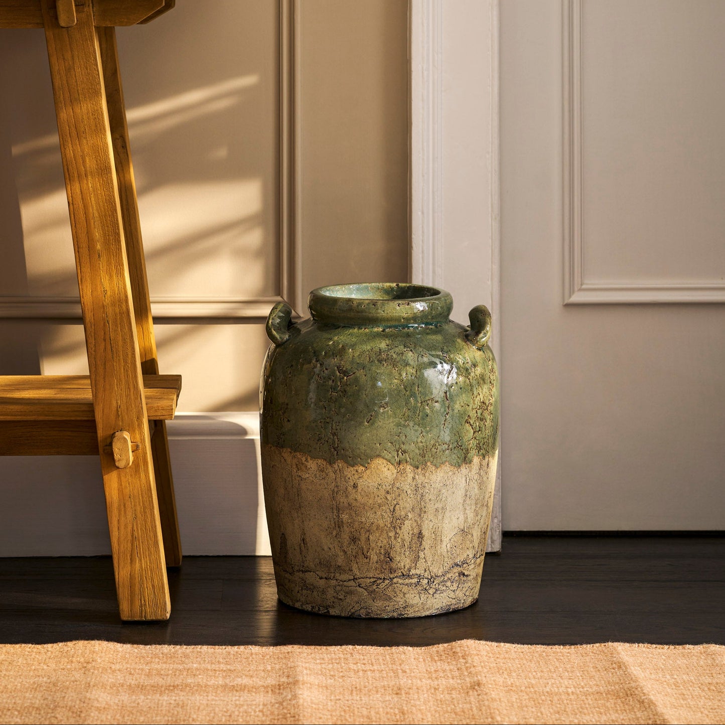Vintage-style green ceramic jar on a dark surface with a wooden stool and neutral wall in the background.